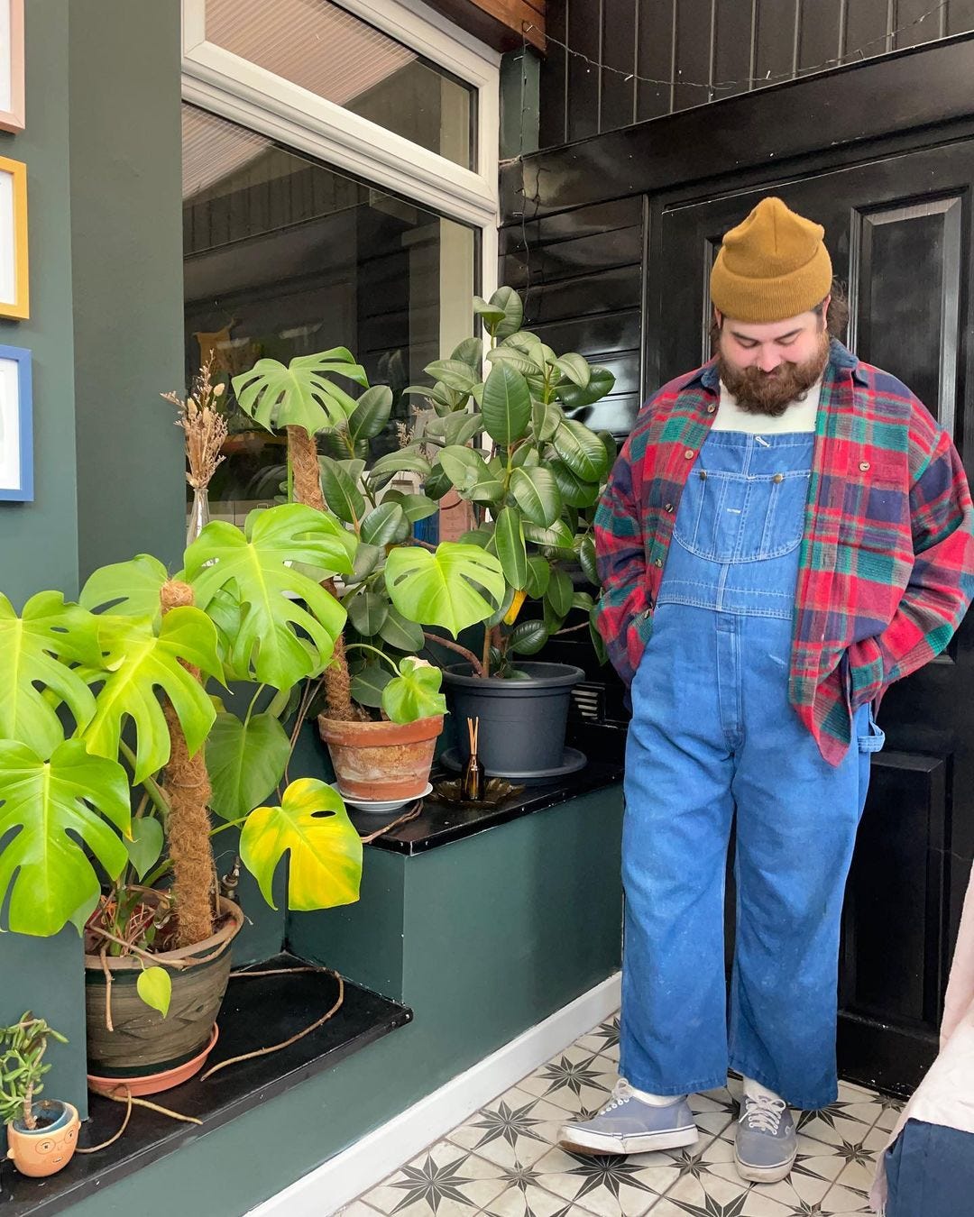 Man standing on porch wearing work clothes with red and green plaid shirt