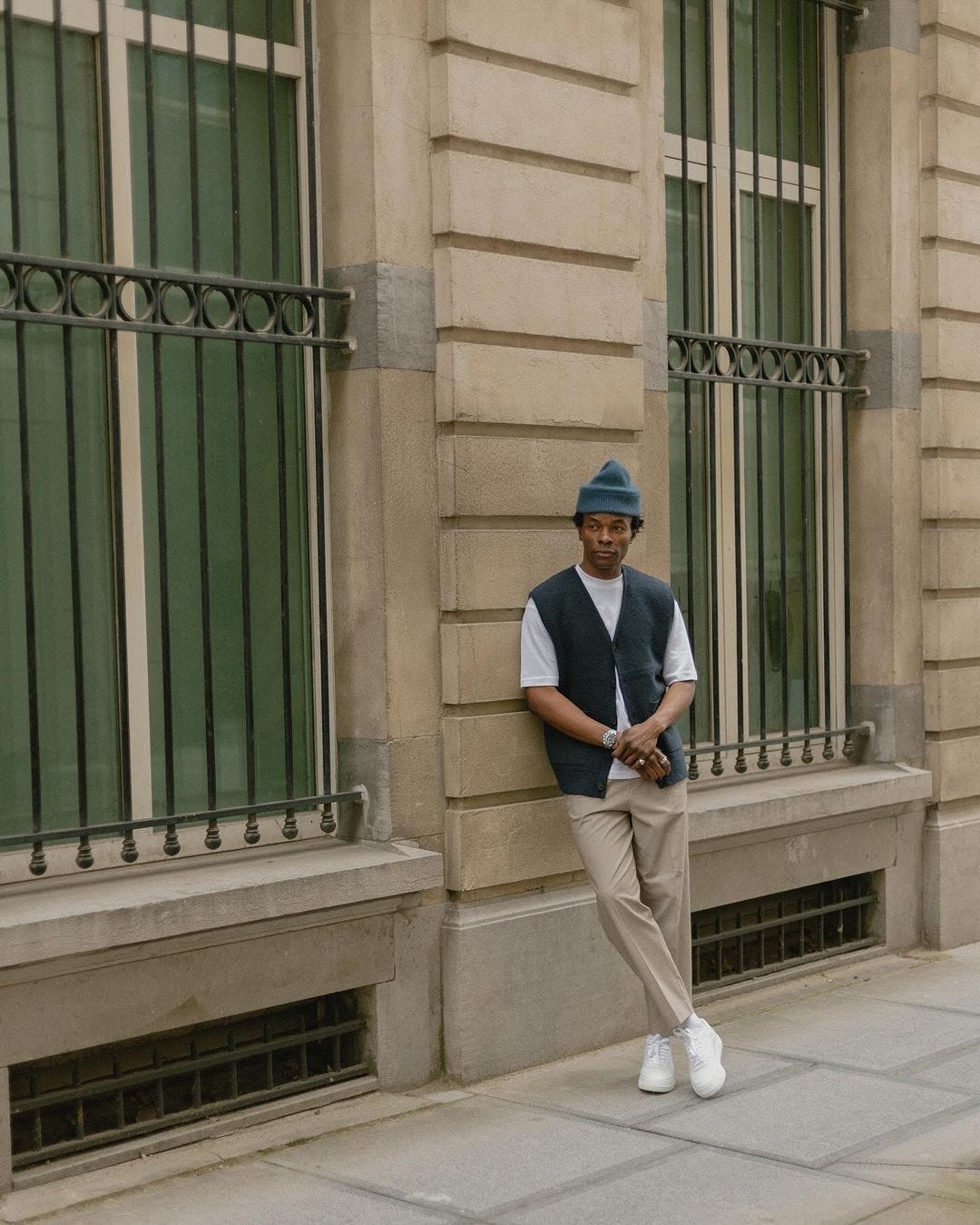 man leaning against a wall wearing a beanie, white t-shirt, dark vest, tan chinos, white socks, and white sneakers