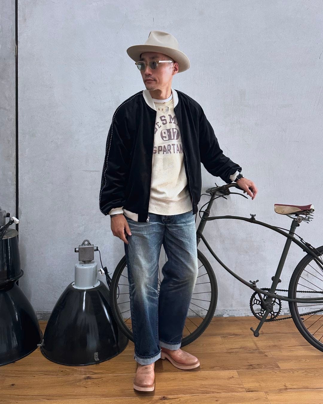 Man standing next to bicycle wearing navy bomber jacket, western hat, sweatshirt and jeans