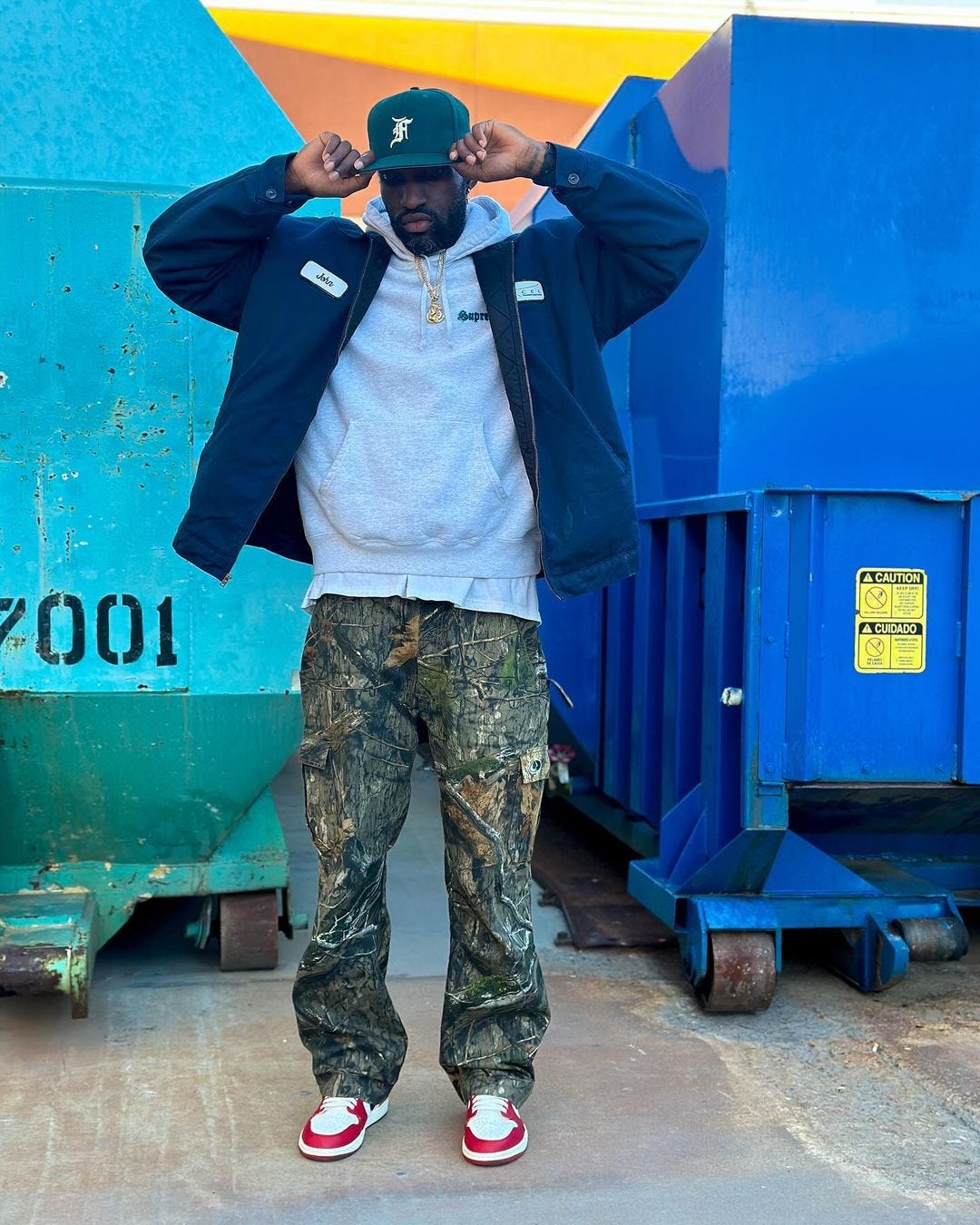 young man standing outside in front of a dumpster wearing a blue workwear jacket, grey hoodie, camo pants and sneakers