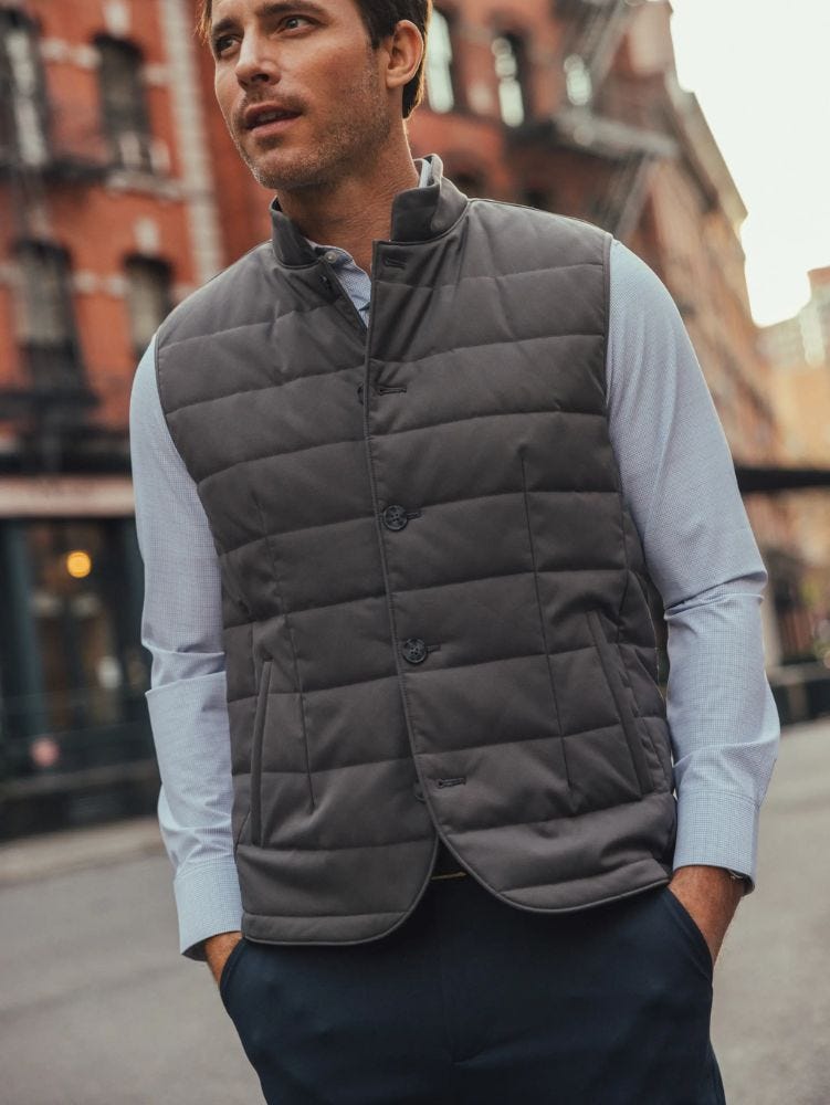 close-up of a man wearing a grey quilted vest over a light blue dress shirt standing outside on the street