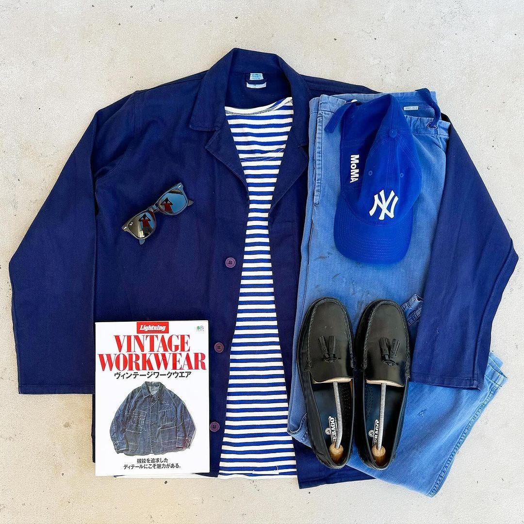 Men's clothing flat lay including blue house coat, blue and white striped shirt, blue baseball cap, blue jeans, black loafers and sunglasses