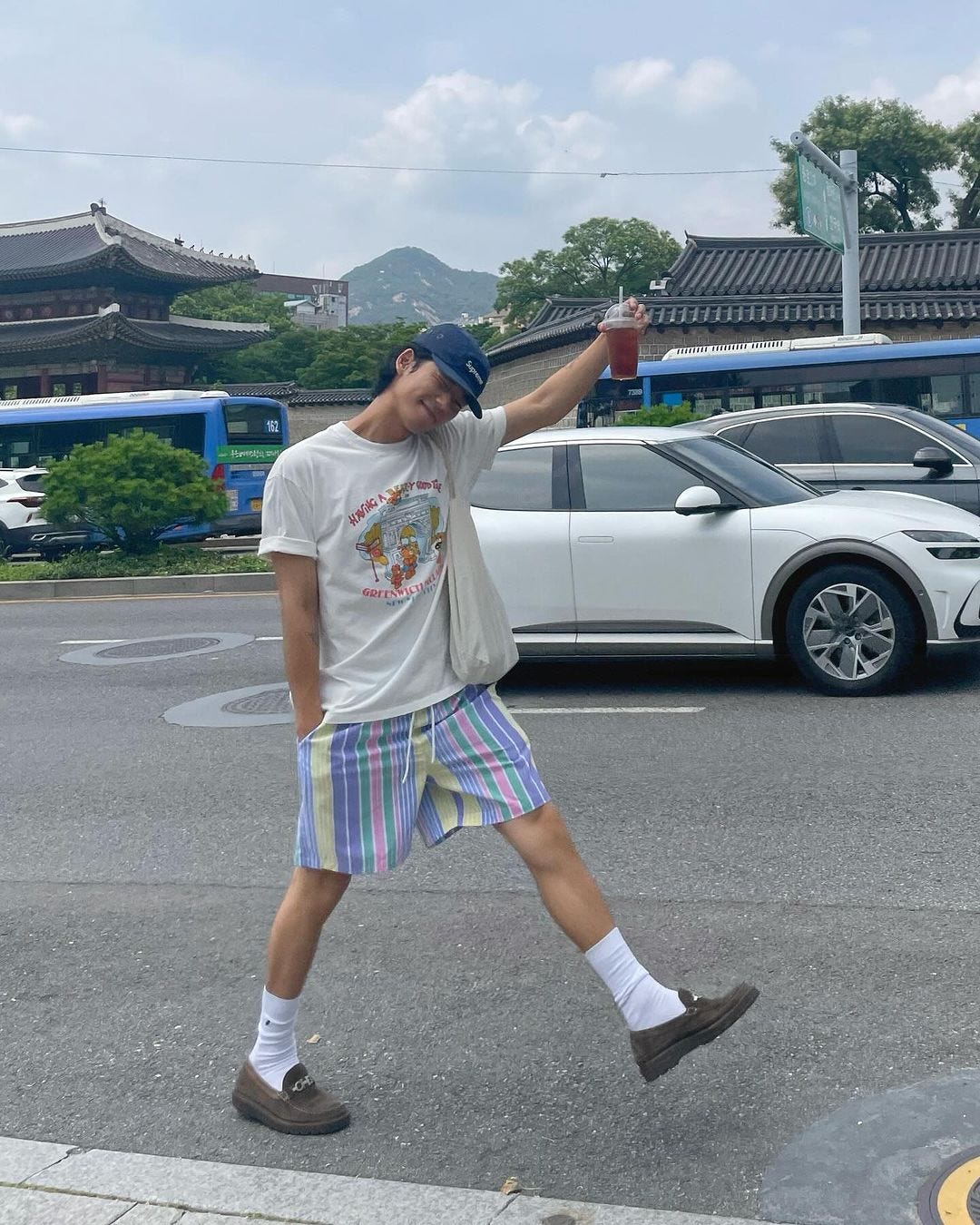 man standing in a parking lot wearing a graphic tee, striped shorts, white socks and loafers, holding up an iced coffee