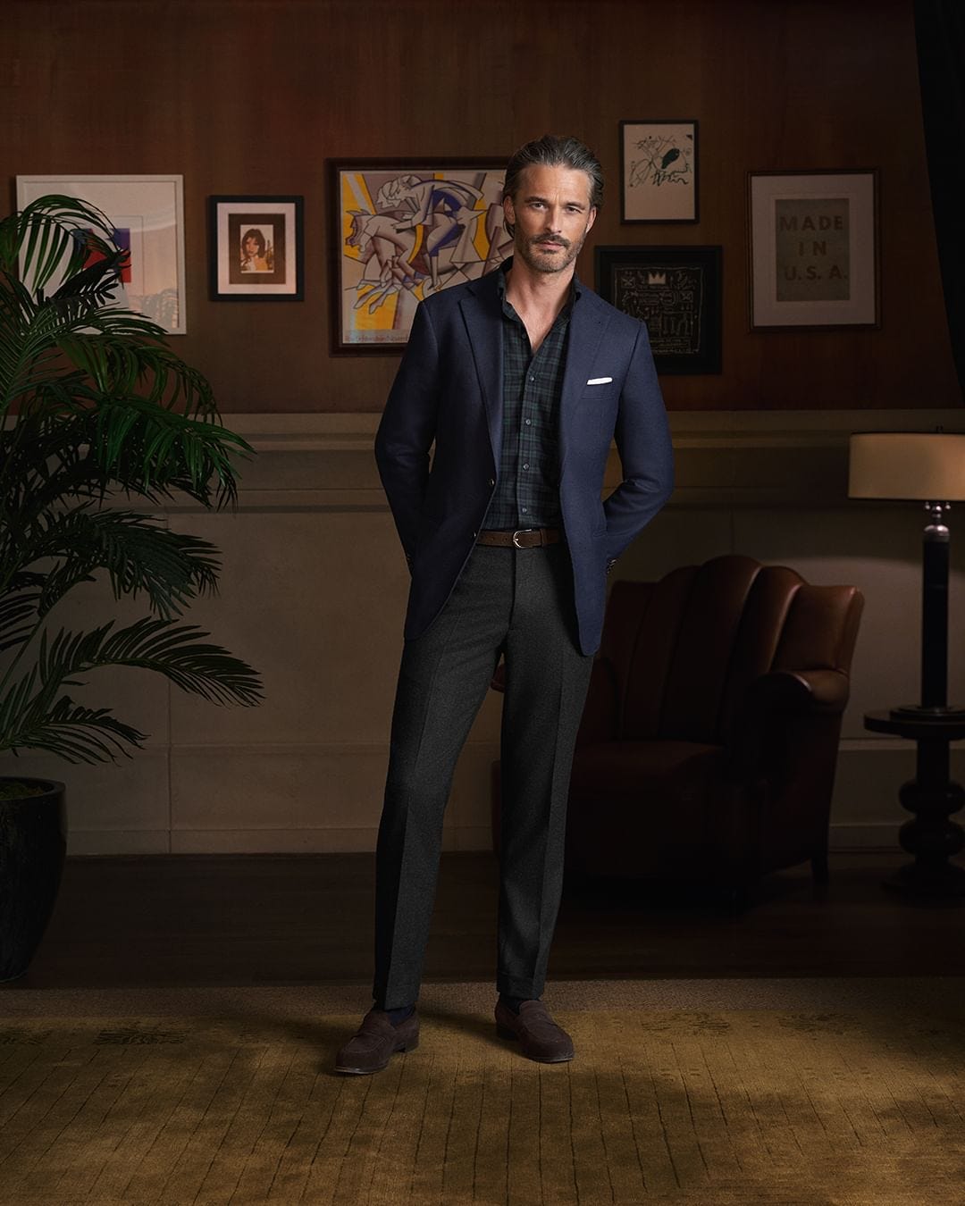 Man in dark study room wearing navy sport coat, plaid shirt and dark pants