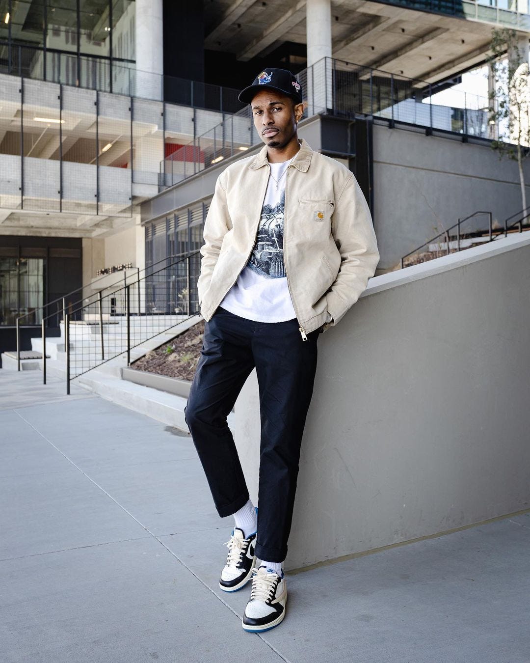 stylish young black man standing outside wearing a cream-colored workwear jacket with dark pants and white sneakers