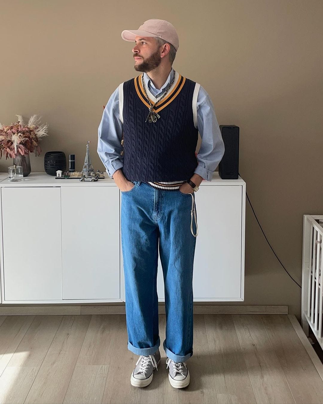 Bearded man wearing navy blue sweater vest with light blue collared shirt, jeans and gray converse sneakers