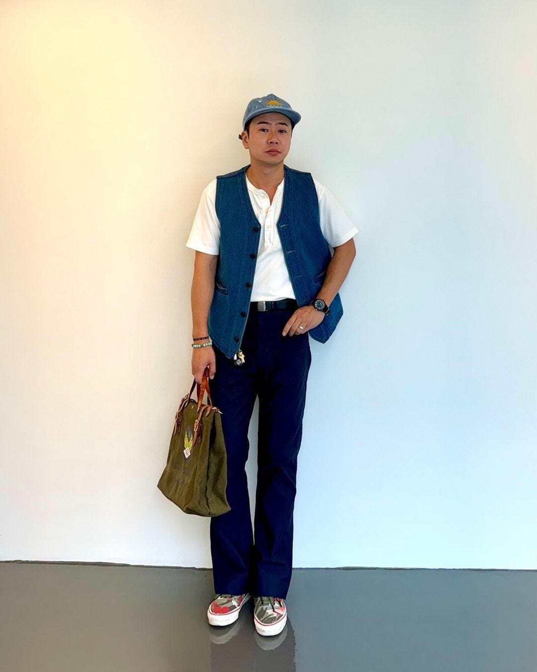 man wearing a baseball cap, white short-sleeve henley shirt, blue denim vest, navy pants  and camo sneakers. he is holding an olive green canvas bag