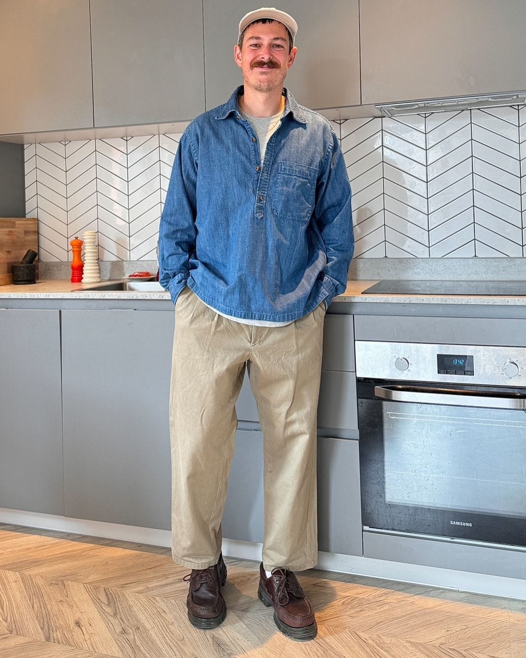 man standing in a kitchen wearing a chambray popover shirt, khaki pants and brown shoes