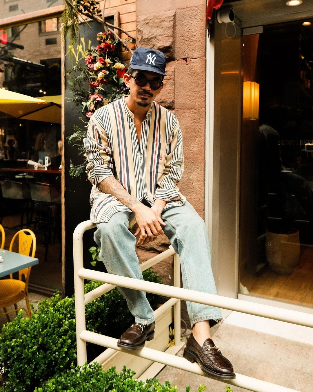 man leaning against a balcony, wearing a blue baseball cap, striped button down shirt, light jeans, and loafers