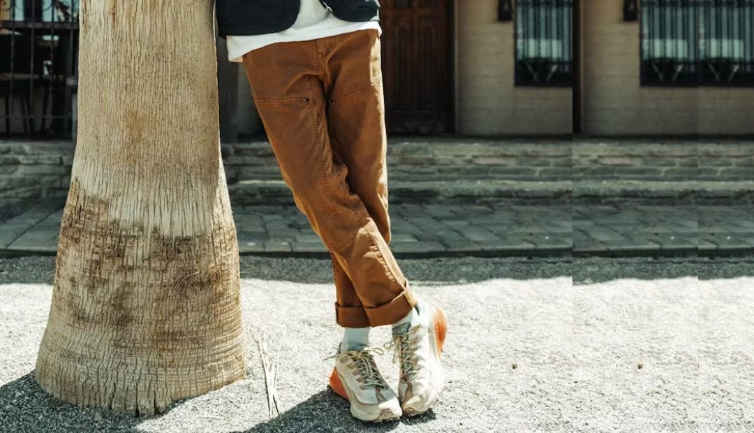 man from the waist down, leaning against a tree and wearing brown workwear pants with grey and orange sneakers