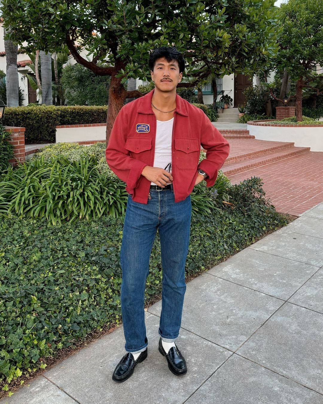 man standing outside wearing a red jacket over a white tank with blue jeans and black loafers