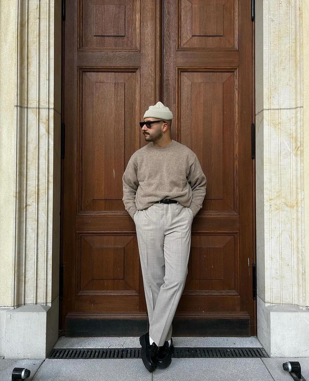 man standing in a doorway wearing a beanie, sweater, chinos, and loafers