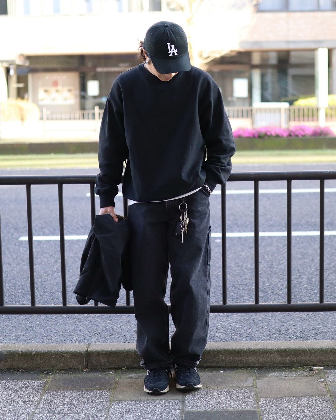 A man stands in front of a barrier wearing a black sweatshirt, black wide pants and black sneakers