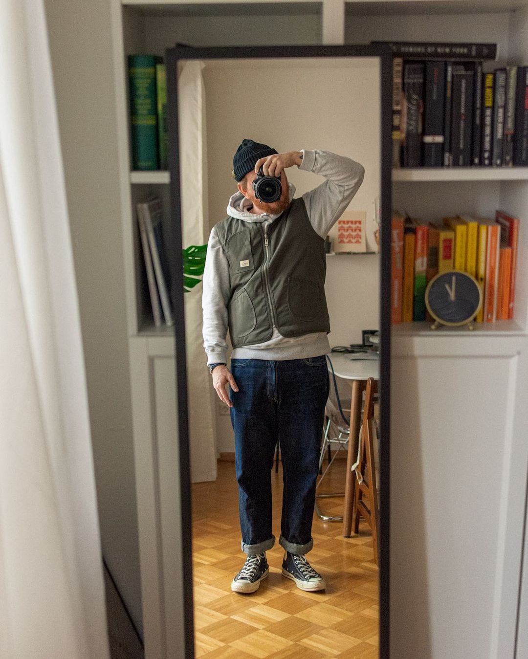 man taking a selfie in a full-length mirror, wearing a beanie, olive green vest over a grey shirt, blue jeans and high top converse sneakers