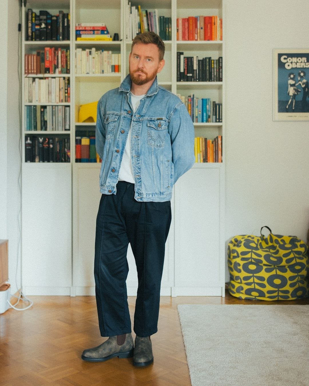 man standing in front of a bookcase with colorful books, wearing a light wash denim jacket unbuttoned over a white t-shirt with navy pants and brown chelsea boots