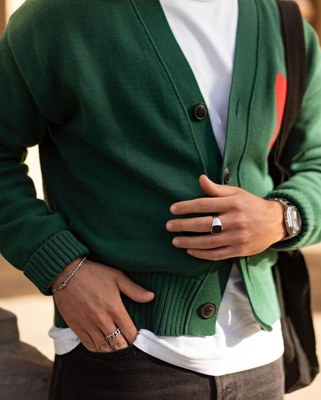 close-up of a man wearing a green cardigan with red heart on the chest, over a white t-shirt, wearing two rings and a bracelet