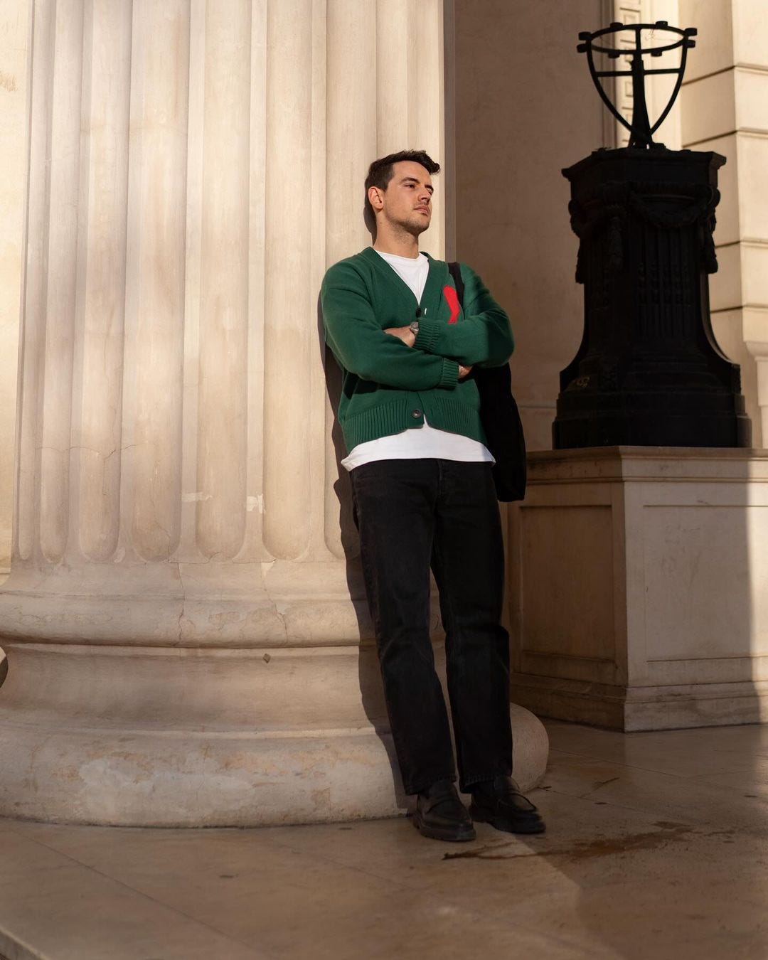 man leaning against a large column, wearing a green cardigan over a white t-shirt with dark pants