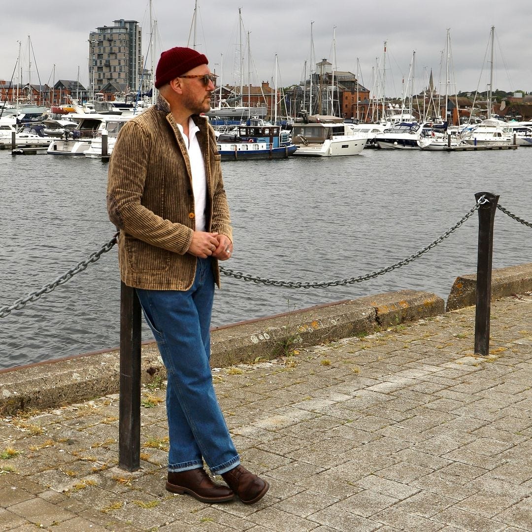 man standing outside in front of boats on the water, wearing a maroon beanie, brown corduroy blazer over a white collared shirt with blue jeans and brown boots