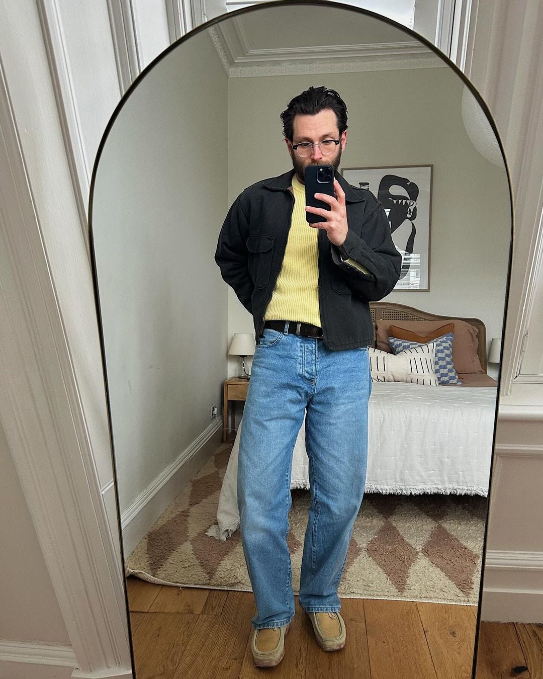 man taking a selfie in a bedroom mirror wearing a yellow sweater, black jacket, and blue jeans