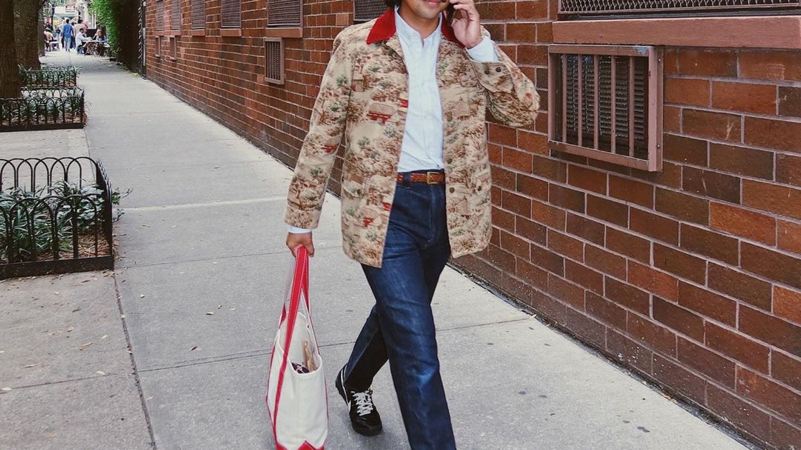 close-up of a man wearing a patterned chore coat with dark blue jeans, carrying a tote bag and talking on the phone