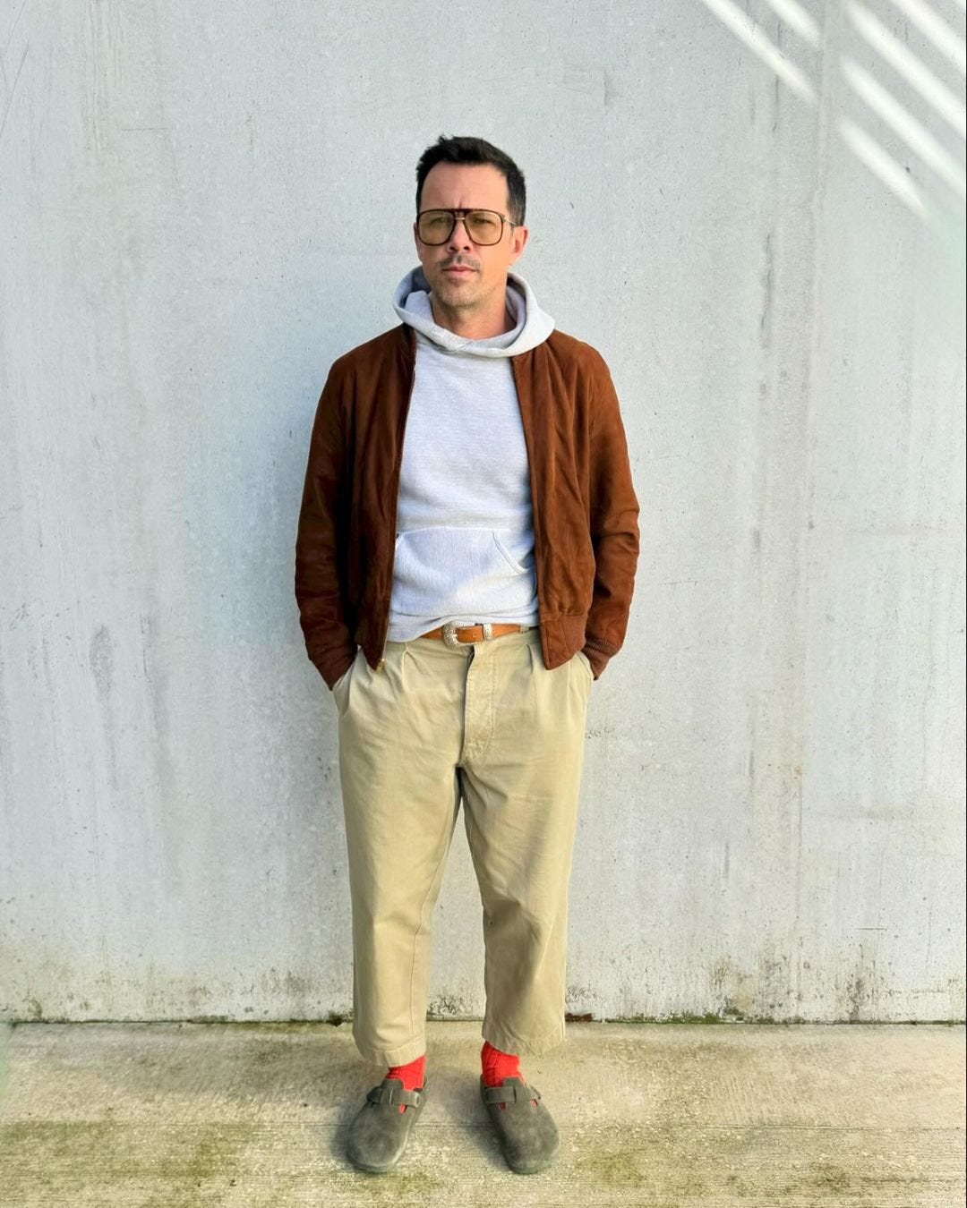 man standing in front of a concrete wall wearing a grey hoodie, brown suede bomber, cropped khaki pants, brown belt, red socks, and suede birkenstock clogs