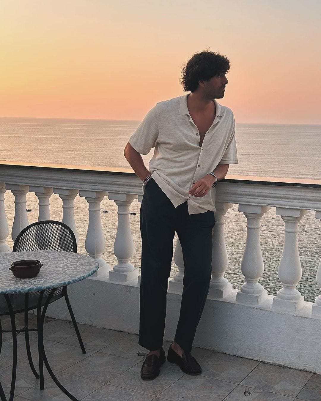 man leaning on a balcony outside, wearing a short-sleeve button down shirt with dark pants and loafers, looking out at the water