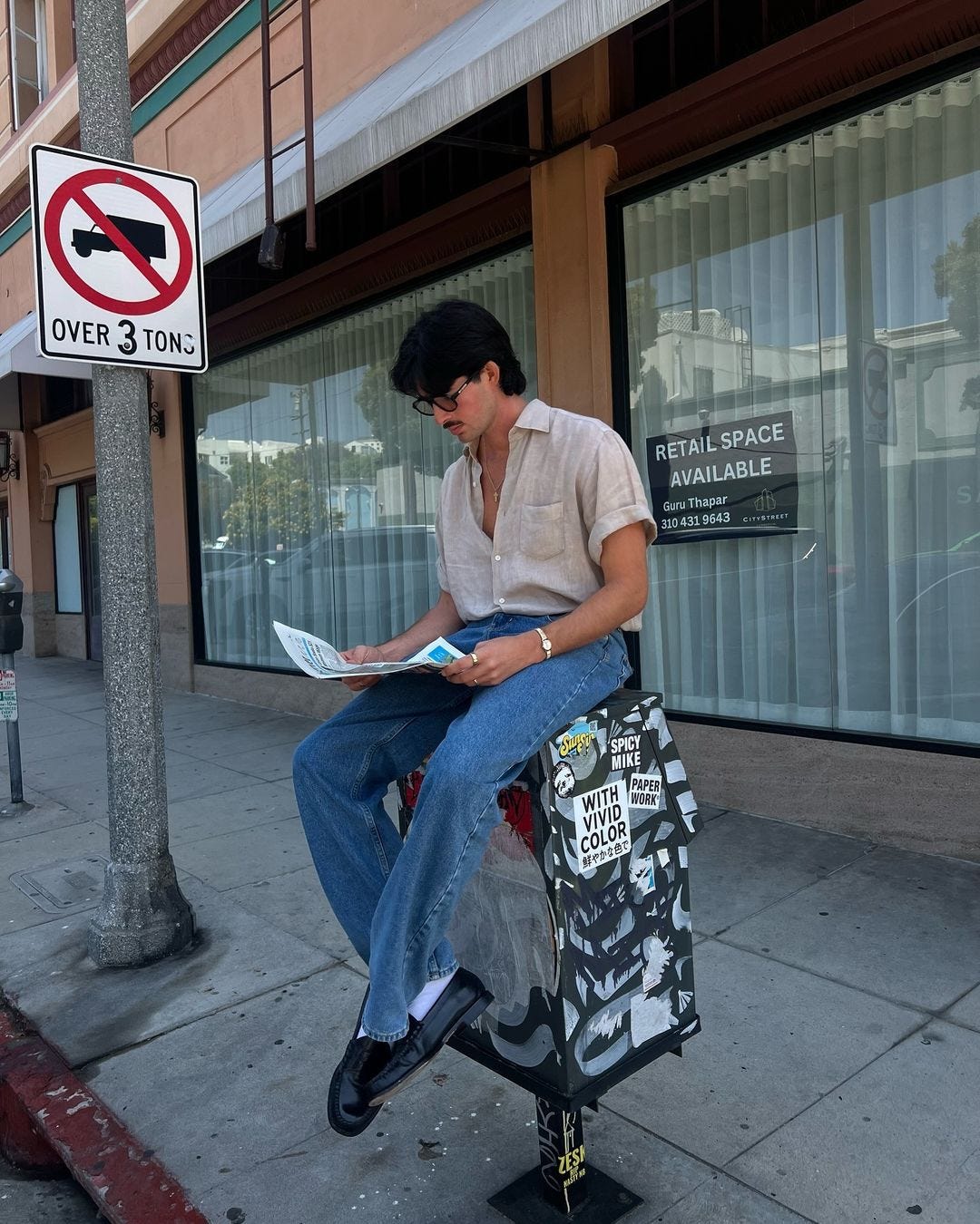 man sitting on a postal box, wearing a short sleeve shirt, jeans, and black loafers