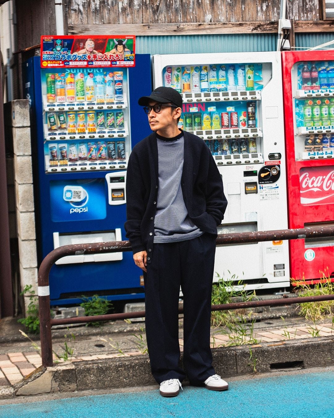 A man stands in front of a row of vending machines, wearing a black woolen sweater, a striped shirt, and loose-cut black trousers