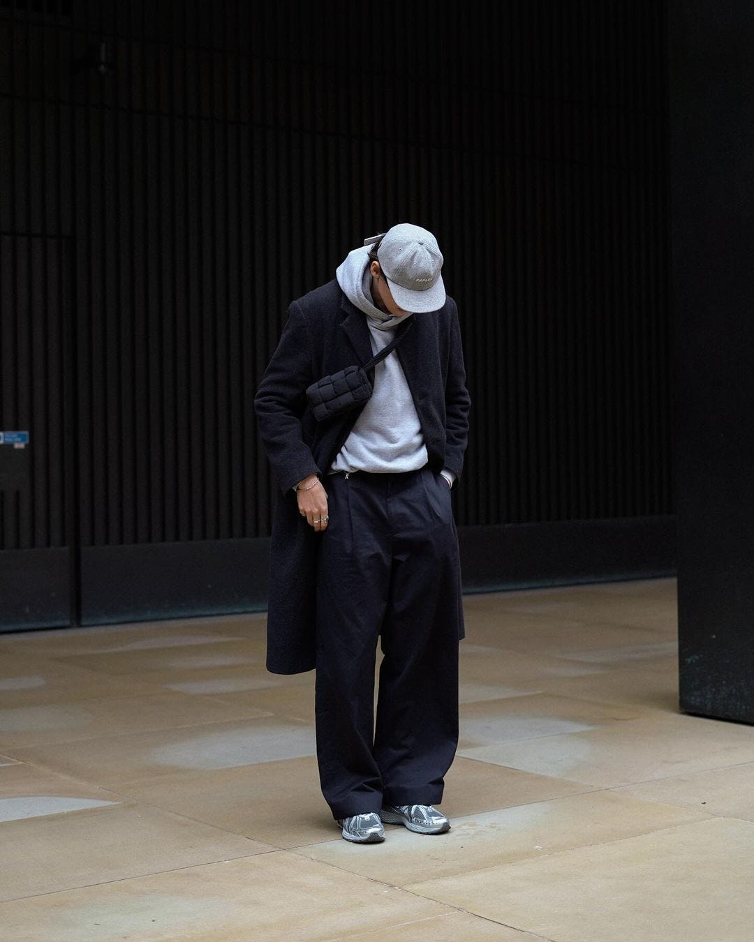 man looking down wearing a grey dad hat, grey hoodie, black topcoat, black crossbody bag, black baggy pants and grey sneakers