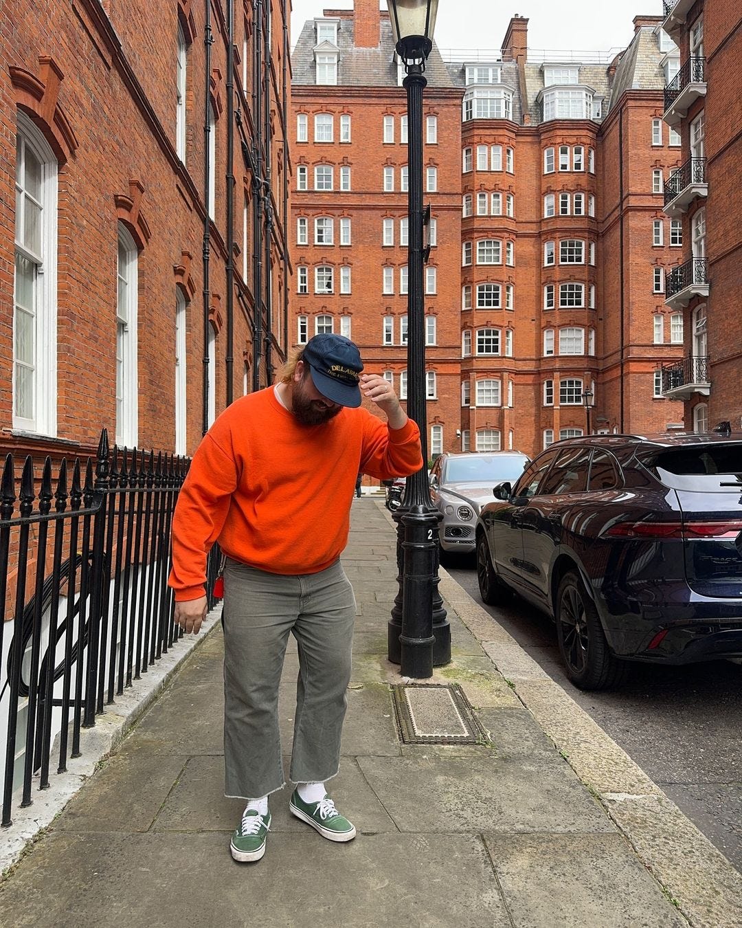 man looking down, standing outside wearing a bright orange sweatshirt, navy baseball cap, grey pants, and green canvas sneakers