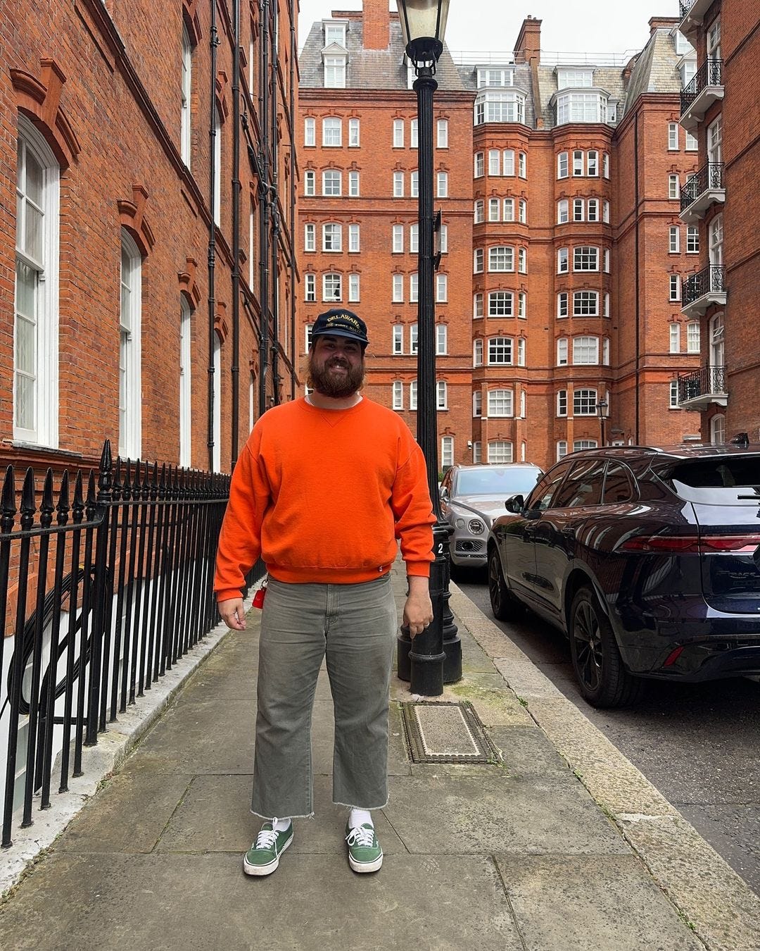 man standing outside wearing a bright orange sweatshirt, navy baseball cap, grey pants, and green canvas sneakers