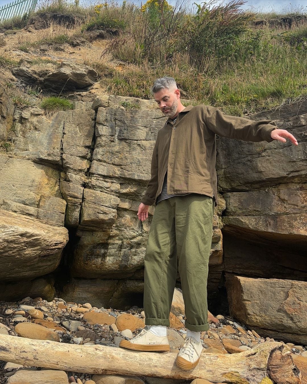 A man balancing on a fallen tree trunk, wearing a brown jacket, olive trousers and cream sneakers with rubber soles