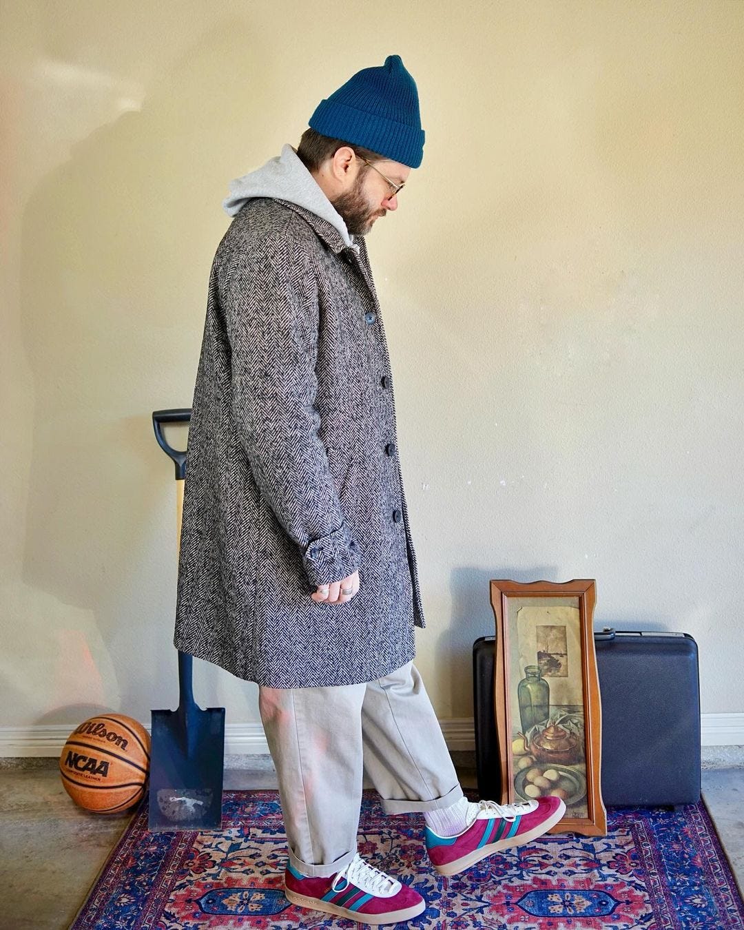 man standing sideways wearing a blue beanie, textured topcoat, grey hoodie, tan khakis, and red and blue sneakers