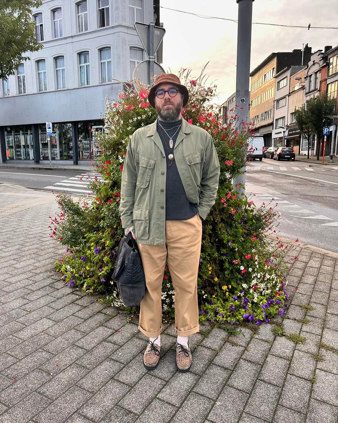 man standing outside on a cobblestone street wearing an olive green jacket with tan chinos, necklaces around his neck, and a bucket hat