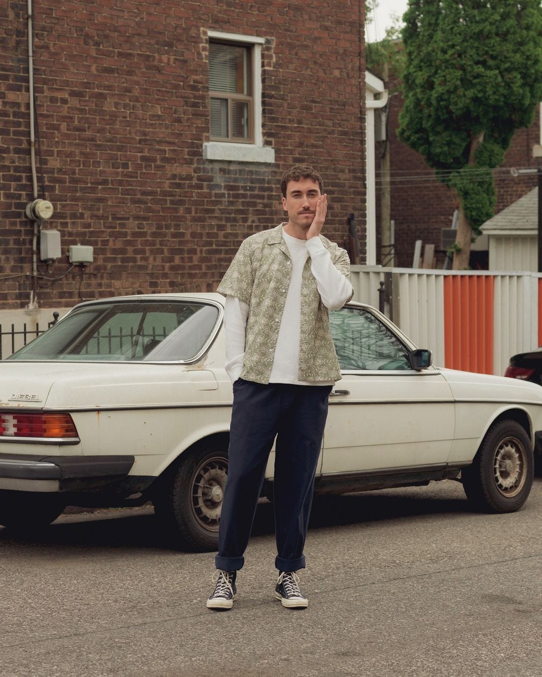 man standing on the street in front of a car wearing a short-sleeve patterned shirt over a long-sleeve shirt with dark pants rolled up at the ankle with black converse all star sneakers