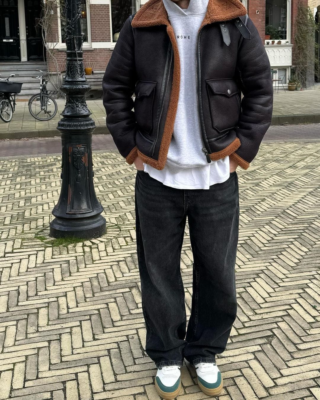 close-up of a man wearing a shearling-lined bomber jacket, grey hoodie, wide-leg black jeans and white gum sole sneakers