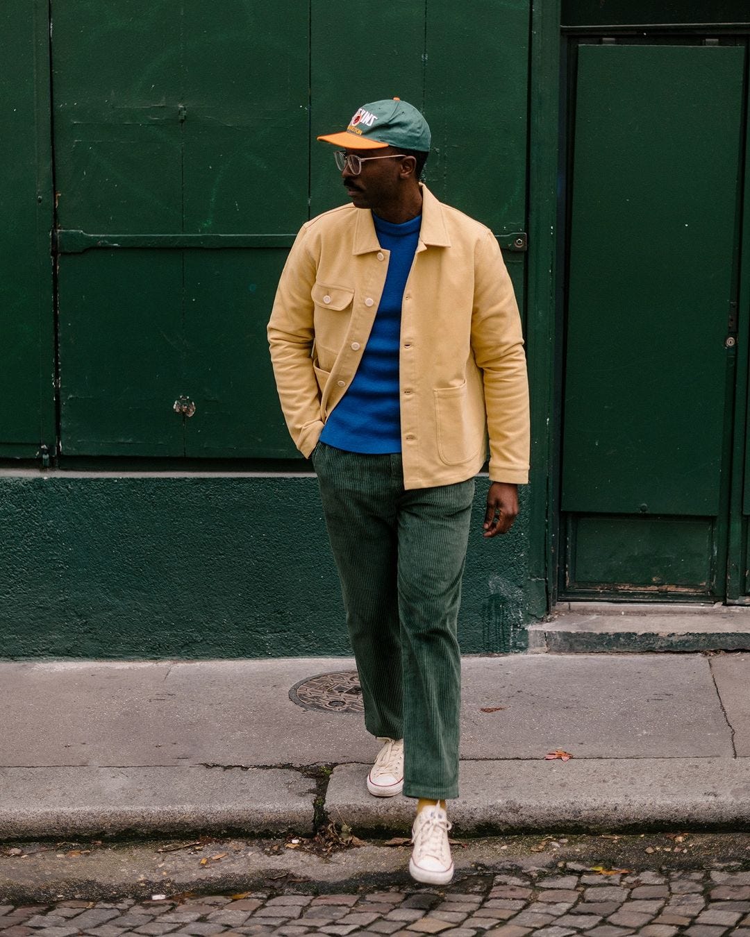 man stepping off a curb wearing a tan chore coat, blue sweater and green corduroy pants