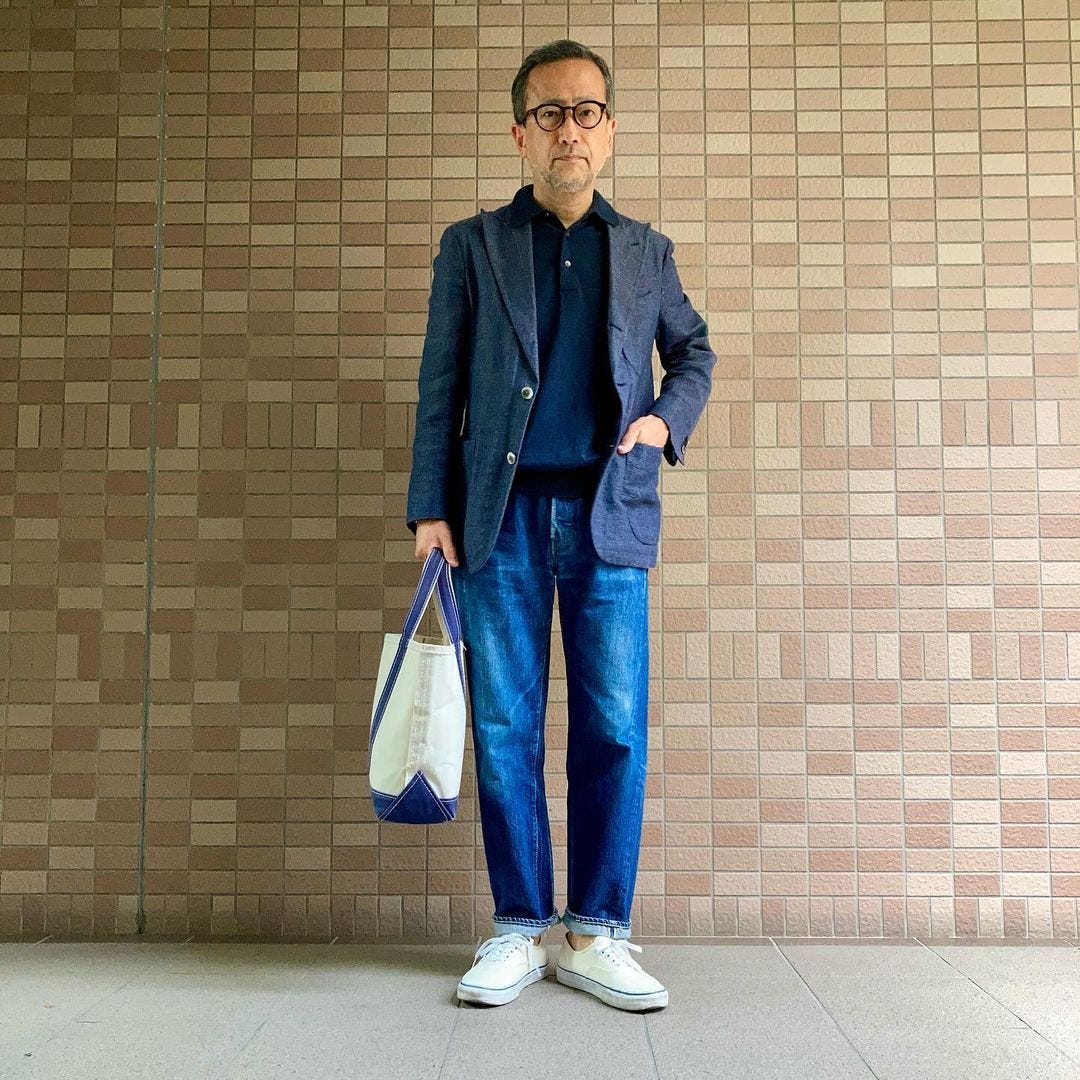 man standing in front of a brick wall wearing a navy blazer over a dark blue knit polo with blue jeans and white sneakers. he's holding a tote bag