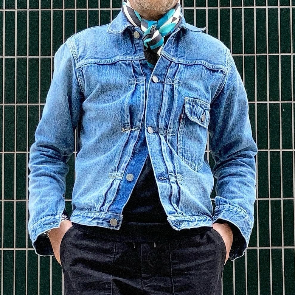 close-up of a man wearing a blue denim jacket buttoned up with a patterned scarf and dark shirt with black pants