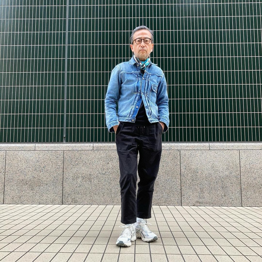 man standing outside wearing a blue denim jacket buttoned up with a patterned scarf and black pants with white sneakers