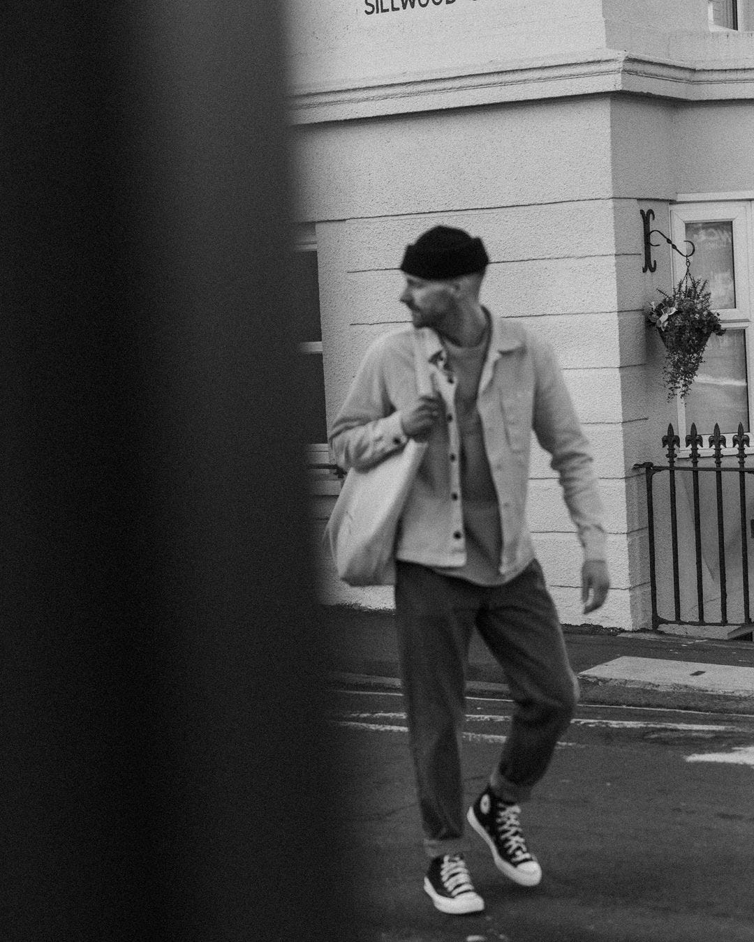 black and white image of a man walking on the street wearing a beanie, overshirt, jeans, and high top sneakers