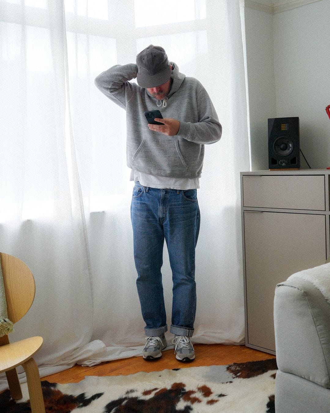 man looking at his phone, wearing a grey baseball cap, grey hoodie, white t-shirt peeking out underneath, blue jeans, and grey new balance sneakers