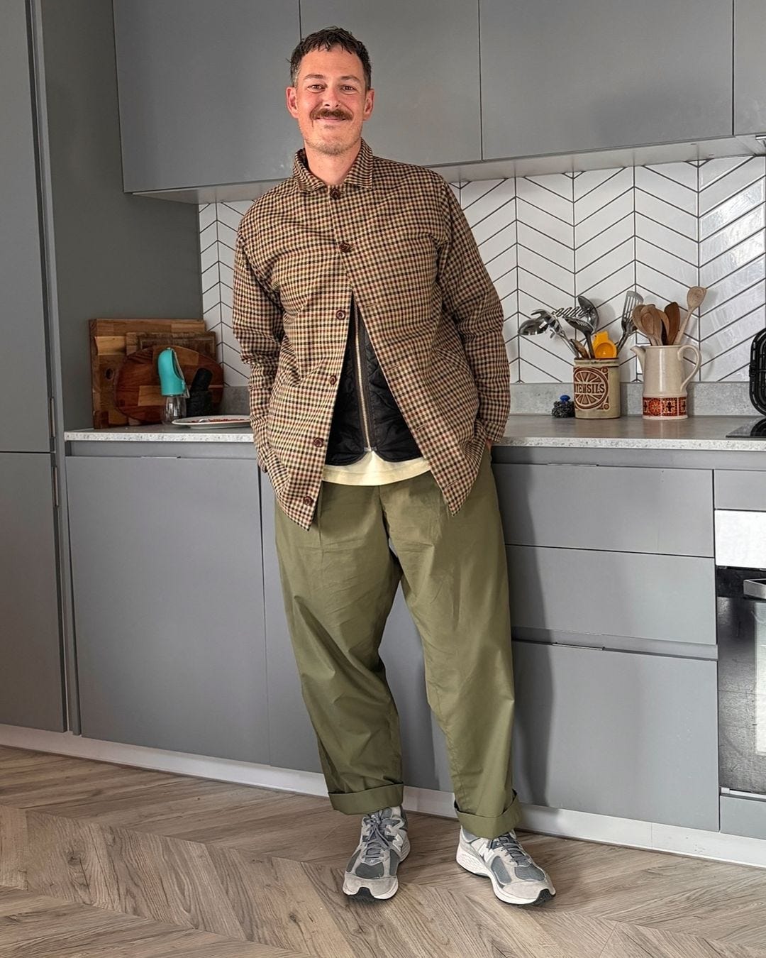 man standing in a kitchen wearing a patterned jacket over a black vest and cream-colored t-shirt with pleated olive green pants and sneakers