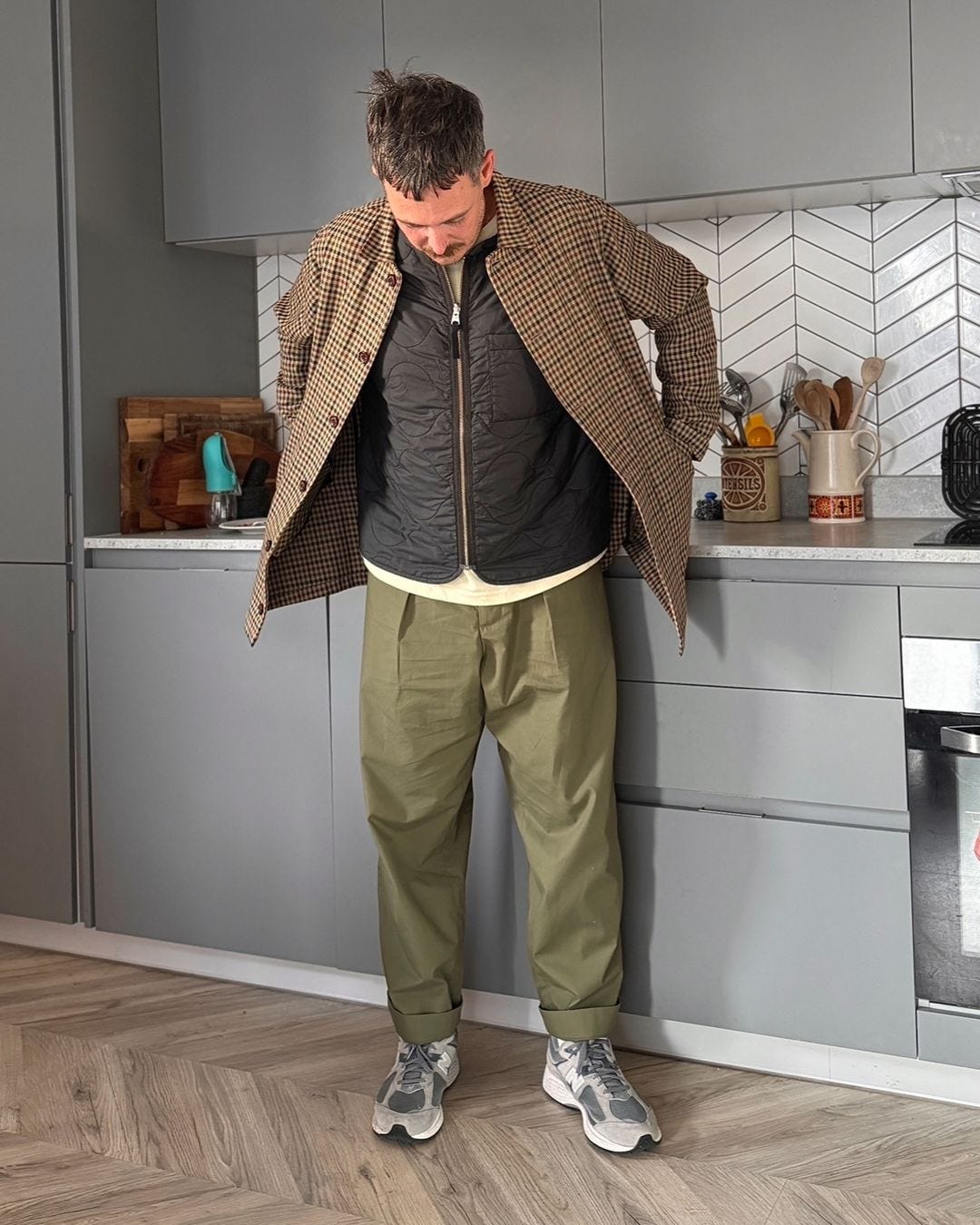 man standing in a kitchen with his hands in his pockets, wearing a patterned jacket over a black vest and cream-colored t-shirt with pleated olive green pants and sneakers