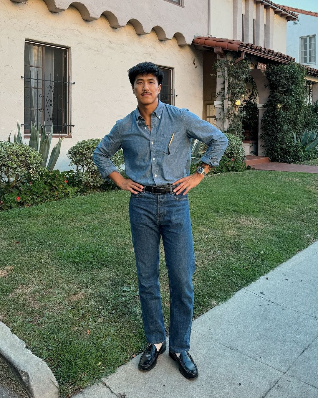 man standing on the front lawn of a house wearing a chambray shirt with jeans and black loafers