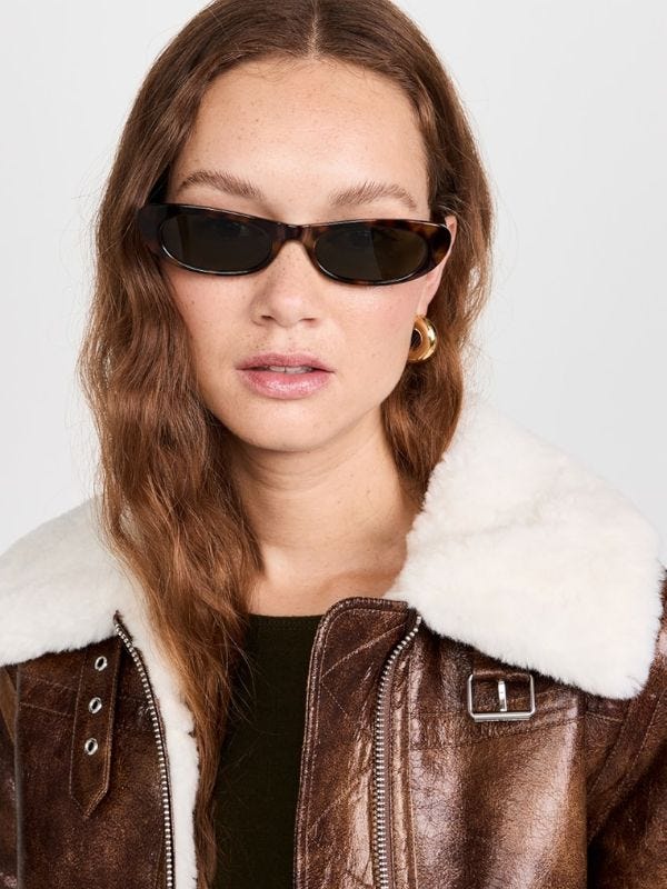 hip woman wearing skinny tortoiseshell sunglasses and a brown leather bomber jacket with gold hoops