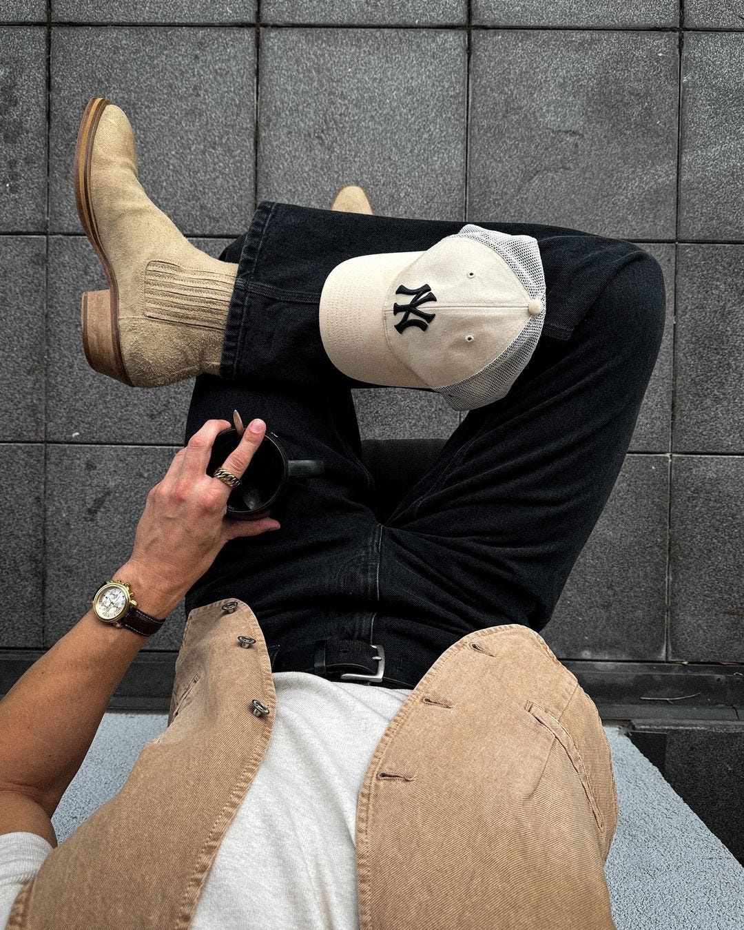 image of a seated man, taken from above, wearing a white t-shirt, tan vest, black jeans, black belt, and tan cowboy boots. a baseball hat is sitting on his knee