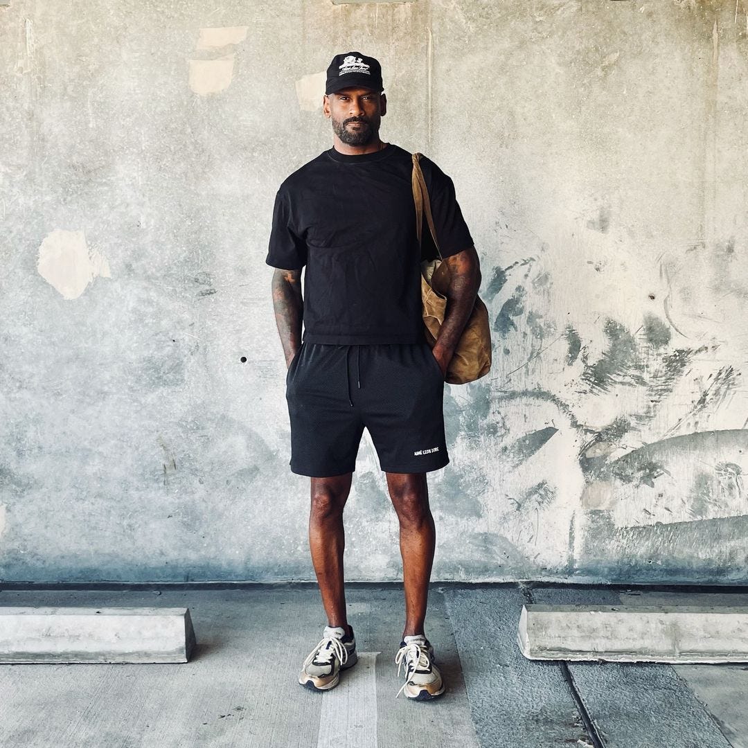 man standing in a parking lot wearing a dark baseball cap, black t-shirt, black shorts, and grey sneakers