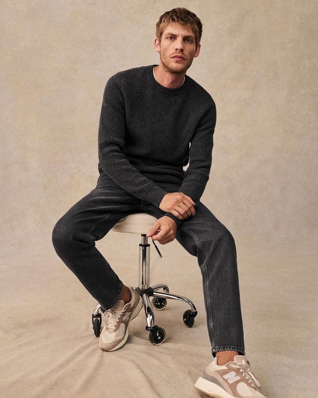 man sitting on a rolling stool wearing a faded black sweater and black jeans