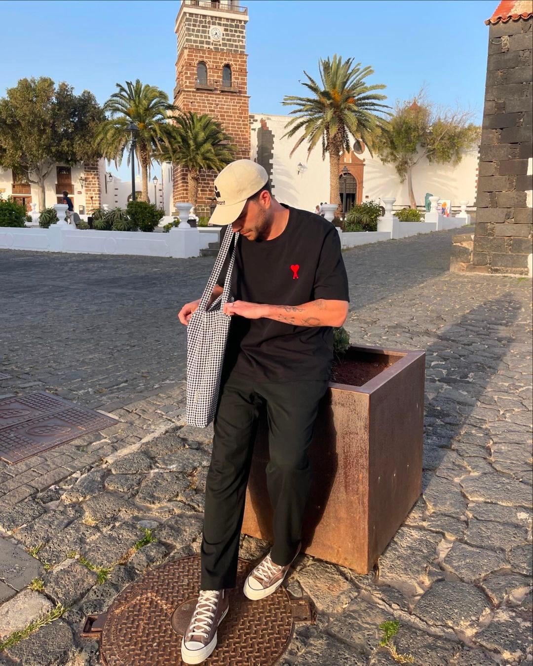 man outside leaning against a bench wearing a black t-shirt with a red logo, black pants and sneakers